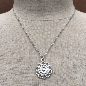 Puede incluir: Collar plateado con un colgante floral detallado. El colgante est&aacute; suspendido de una fina cadena. El collar se muestra sobre un fondo de tono neutro.
