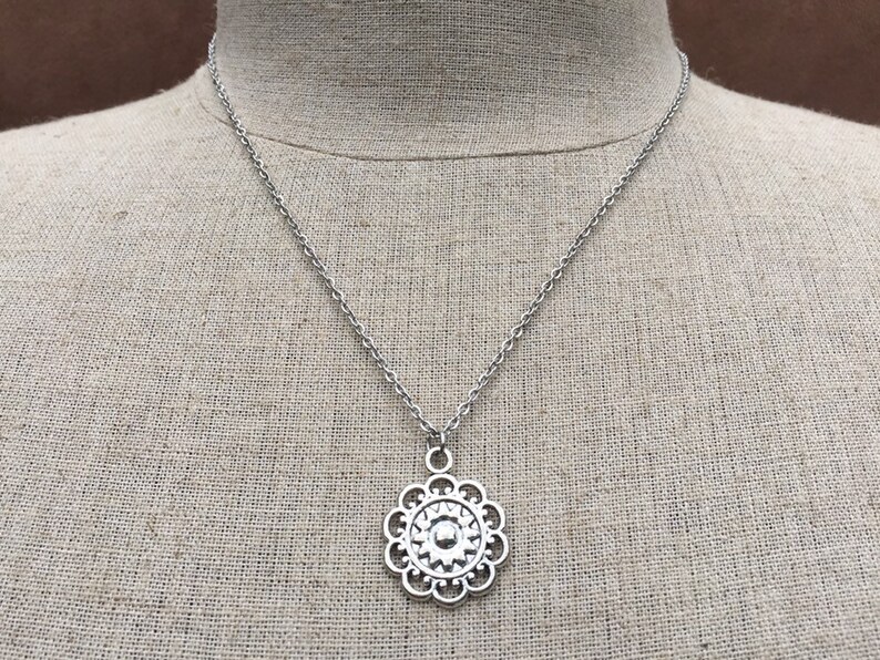Puede incluir: Collar plateado con un colgante floral detallado. El colgante est&aacute; suspendido de una fina cadena. El collar se muestra sobre un fondo de tono neutro.