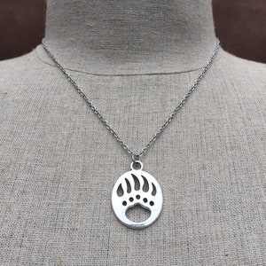May include: A silver chain necklace with a pendant shaped like a bear paw print. The pendant is oval and has a cut-out design.