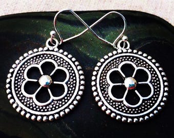 May include: A pair of silver-colored earrings. Each earring features a circular design with a floral pattern in the center and a beaded border. The earrings have a hook closure.