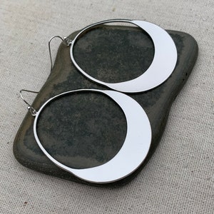 Puede incluir: Un par de pendientes de aro grandes de color plateado. Cada pendiente presenta un dise&ntilde;o en forma de media luna dentro del marco circular. Los pendientes se muestran sobre una piedra gris oscura sobre un fondo neutro.