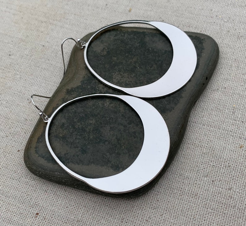 Puede incluir: Un par de pendientes de aro grandes de color plateado. Cada pendiente presenta un dise&ntilde;o en forma de media luna dentro del marco circular. Los pendientes se muestran sobre una piedra gris oscura sobre un fondo neutro.