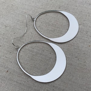 Puede incluir: Un par de pendientes de tono plateado con forma abierta y asim&eacute;trica de l&aacute;grima o media luna. Cada pendiente cuelga de un fino gancho de metal curvado y presenta una superficie met&aacute;lica lisa y reflectante con acabado pulido.