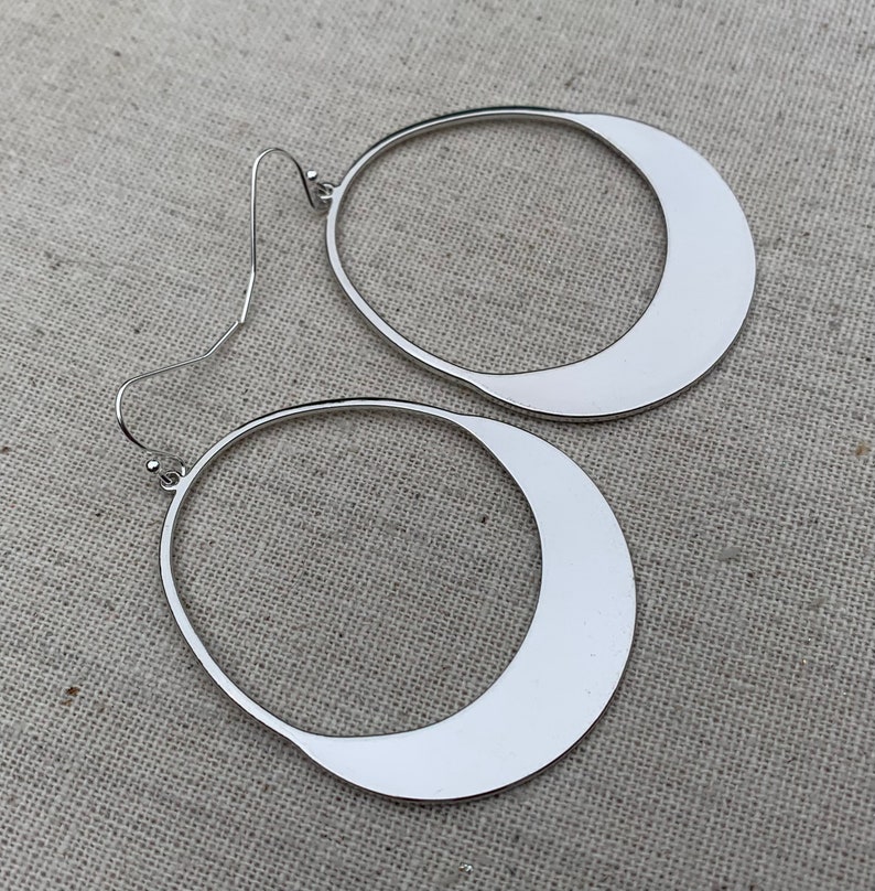 Puede incluir: Un par de pendientes de tono plateado con forma abierta y asim&eacute;trica de l&aacute;grima o media luna. Cada pendiente cuelga de un fino gancho de metal curvado y presenta una superficie met&aacute;lica lisa y reflectante con acabado pulido.