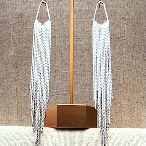 Può includere: Un paio di lunghi orecchini pendenti color argento caratterizzati da una montatura superiore triangolare geometrica collegata a una frangia a cascata di sottili catenelle metalliche che pendono verticalmente per un effetto scintillante.