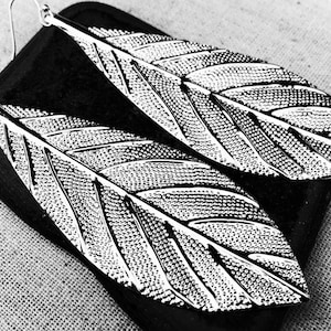 May include: A pair of silver-colored feather earrings. The earrings are detailed with intricate veining and texture, resembling real feathers. The earrings are displayed on a dark, textured surface, set against a neutral background.