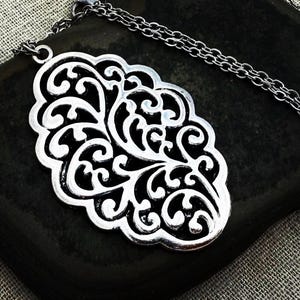 May include: Silver-toned filigree pendant necklace. The pendant has an ornate, openwork design with swirling patterns. The necklace chain is a delicate, linked style. The pendant is displayed on a dark, textured surface.