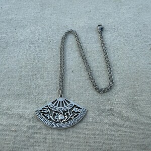 May include: Silver-toned pendant necklace featuring a fan-shaped charm with intricate floral and geometric designs. The pendant hangs from a delicate chain. The necklace is displayed on a neutral linen surface.