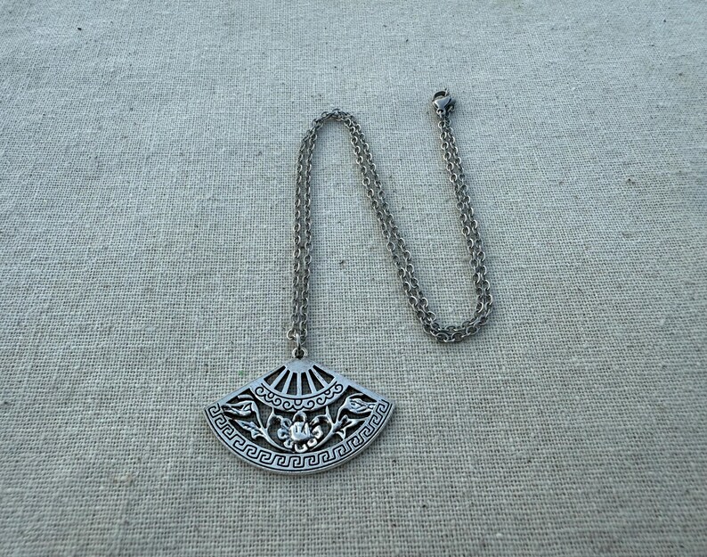 May include: Silver-toned pendant necklace featuring a fan-shaped charm with intricate floral and geometric designs. The pendant hangs from a delicate chain. The necklace is displayed on a neutral linen surface.