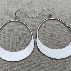 Puede incluir: Un par de pendientes de aro plateados. Cada pendiente presenta un dise&ntilde;o circular abierto grande con una base plana en forma de media luna. Los pendientes tienen una est&eacute;tica sencilla y moderna, adecuada para cualquier ocasi&oacute;n.