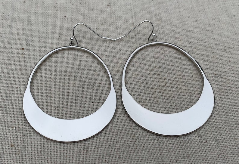Puede incluir: Un par de pendientes de aro plateados. Cada pendiente presenta un dise&ntilde;o circular abierto grande con una base plana en forma de media luna. Los pendientes tienen una est&eacute;tica sencilla y moderna, adecuada para cualquier ocasi&oacute;n.