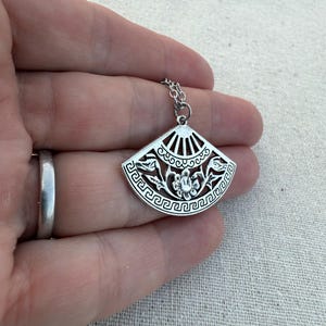 May include: Silver-toned pendant necklace featuring a fan-shaped charm. The charm has an intricate design with floral and geometric patterns. The necklace is displayed on a hand, showcasing the delicate details of the jewelry.