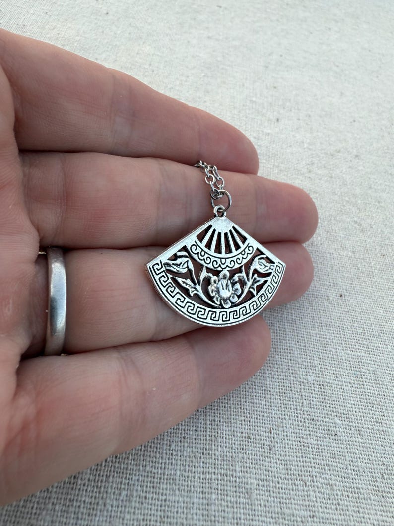 May include: Silver-toned pendant necklace featuring a fan-shaped charm. The charm has an intricate design with floral and geometric patterns. The necklace is displayed on a hand, showcasing the delicate details of the jewelry.