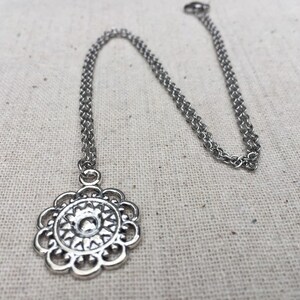 Puede incluir: Un collar plateado con un colgante floral. El colgante presenta un dise&ntilde;o calado detallado con un elemento central en relieve. La cadena es de estilo fino y enlazado. El collar se muestra sobre un fondo neutro y texturizado.