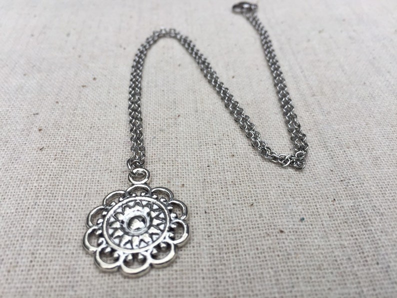 Puede incluir: Un collar plateado con un colgante floral. El colgante presenta un dise&ntilde;o calado detallado con un elemento central en relieve. La cadena es de estilo fino y enlazado. El collar se muestra sobre un fondo neutro y texturizado.