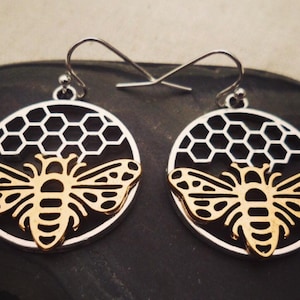 May include: A pair of silver-tone bee earrings. Each earring features a gold-colored bee design set within a silver-tone circle with a honeycomb pattern. The earrings have a hook closure.