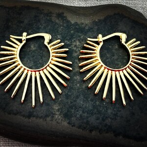 May include: A pair of gold-colored hoop earrings. Each earring features a sunburst design with radiating spikes. The earrings are displayed on a dark, textured surface, showcasing their bright, metallic finish.