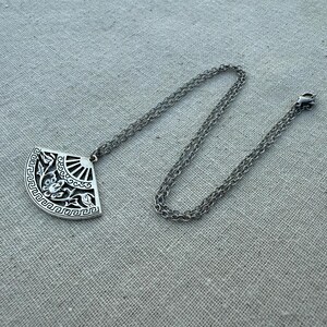 May include: A silver-toned pendant necklace featuring a fan-shaped pendant with intricate floral and geometric designs. The pendant is suspended from a delicate chain with a lobster clasp. The necklace is displayed on a neutral linen surface.