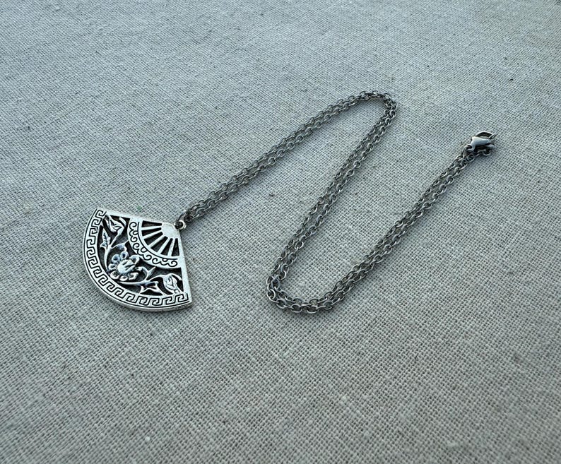 May include: A silver-toned pendant necklace featuring a fan-shaped pendant with intricate floral and geometric designs. The pendant is suspended from a delicate chain with a lobster clasp. The necklace is displayed on a neutral linen surface.