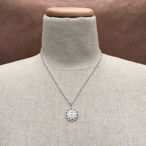 Puede incluir: Collar plateado con un colgante floral. El colgante presenta un dise&ntilde;o calado detallado con un elemento circular central rodeado de formas de p&eacute;talos. El collar se muestra en un maniqu&iacute;.