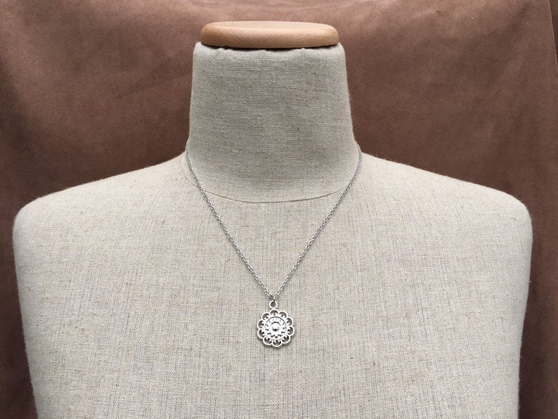 Puede incluir: Collar plateado con un colgante floral. El colgante presenta un dise&ntilde;o calado detallado con un elemento circular central rodeado de formas de p&eacute;talos. El collar se muestra en un maniqu&iacute;.