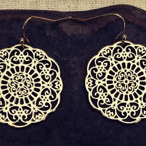 May include: A pair of gold-colored filigree earrings. Each earring features a circular, openwork design with intricate patterns. The earrings have a hook closure and are displayed on a dark, textured surface.