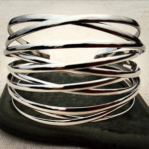 Op de afbeelding: Een brede armband van gepolijst zilverkleurig metaal, bestaande uit meerdere dunne, gladde banden die elkaar overlappen en kruisen in een doorlopend, gelaagd ontwerp, wat een open, kooi-achtige structuur vormt met een reflecterende afwerking.