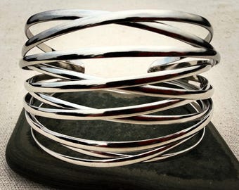 Puede incluir: Un brazalete ancho hecho de metal pulido en tono plateado, con múltiples bandas delgadas y lisas que se superponen y se entrecruzan en un diseño continuo y estratificado, creando una estructura abierta tipo jaula con acabado metálico.