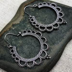 May include: A pair of silver-colored hoop earrings with an intricate, openwork design. The earrings feature a scalloped edge with small circular details and a hook closure. The earrings are displayed on a dark green stone.