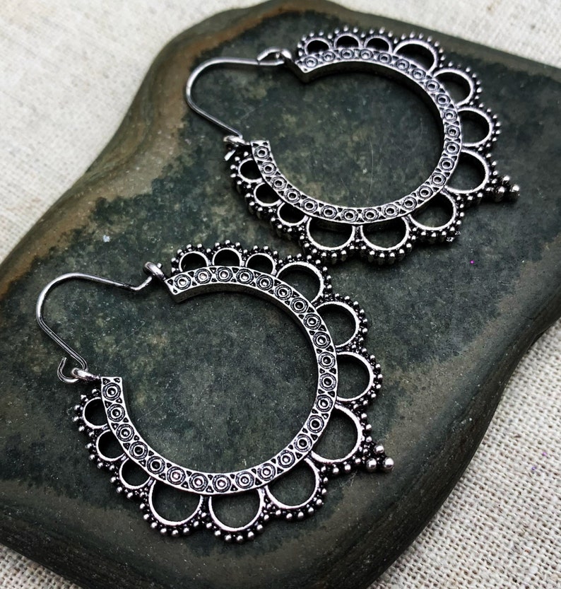 May include: A pair of silver-colored hoop earrings with an intricate, openwork design. The earrings feature a scalloped edge with small circular details and a hook closure. The earrings are displayed on a dark green stone.
