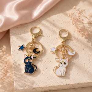 May include: Two metal keychains featuring cat charms with crescent moon symbols on their foreheads. One keychain has a dark blue cat with a blue star and black moon charm, while the other has a white cat with a white star and white moon charm.