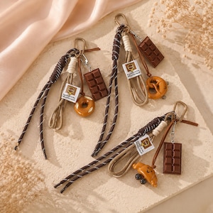 May include: Three keychains featuring braided brown and white cords with metal clips. Each includes a small white tag with a circular logo and text, a brown chocolate bar charm, and a food-shaped charm: a donut, a pretzel, and a croissant.