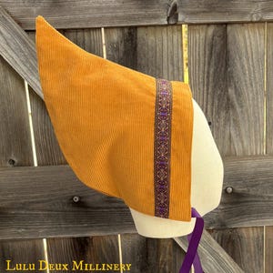May include: A mustard yellow corduroy bonnet featuring a pointed crown and a decorative trim band with a purple and gold geometric pattern. The headwear includes a purple ribbon tie and is displayed on a mannequin head, with the text Lulu Deux Millinery.