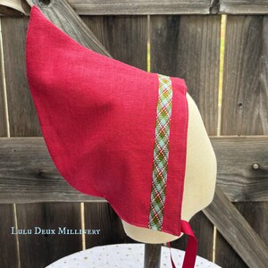 May include: A bright red linen bonnet featuring a pointed crown and a decorative woven trim band with a diamond pattern in green, blue, and white. The bonnet includes matching red fabric ties at the base and is displayed on a mannequin head.