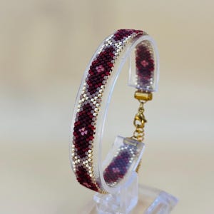 Puede incluir: Una pulsera de cuentas con un patrón geométrico en burdeos, blanco y dorado. La pulsera se exhibe en un soporte de acrílico transparente, mostrando su intrincado diseño. El cierre es de color dorado.
