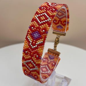 Op de afbeelding: Een kralenarmband met een geometrisch patroon in tinten rood, oranje en paars. De armband wordt tentoongesteld op een transparante standaard, die het ingewikkelde ontwerp en de metalen accenten benadrukt. De sluiting van de armband is goudkleurig.