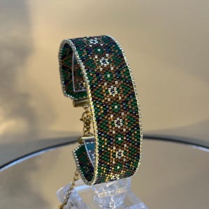 Könnte beinhalten: Ein Armband mit Perlen in einem geometrischen Muster in Grün-, Braun- und Goldtönen. Das Armband wird auf einem transparenten Ständer präsentiert, der sein aufwendiges Design und seine Handwerkskunst hervorhebt. Es ist mit einer goldfarbenen Kette gesichert.