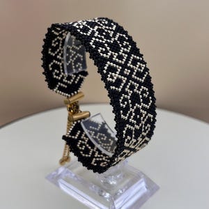 Op de afbeelding: Zwart-witte geweven armband met een geometrisch patroon. De armband wordt gepresenteerd op een transparante standaard met goudkleurige hardware. Het gedetailleerde patroon heeft een herhalend patroon van witte vormen op een zwarte achtergrond.