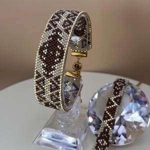 Puede incluir: Un brazalete de cuentas con un patrón geométrico en marrón, blanco y dorado. El brazalete se exhibe sobre un soporte transparente, con un segundo brazalete y un cristal en el fondo. El brazalete mide aproximadamente 3,8 cm de ancho.