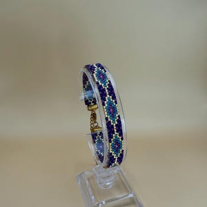 Può includere: Un braccialetto di perline con un motivo geometrico nei toni del blu, verde acqua e oro. Il braccialetto è esposto su un supporto trasparente, che mette in mostra il suo design e la sua lavorazione intricati. Il braccialetto è largo circa 2 cm.