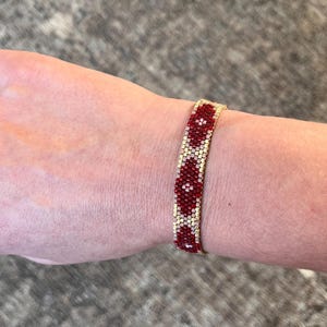 May include: A beaded bracelet with a repeating pattern of red and cream-colored beads. The bracelet is worn on a wrist and has a gold-colored band. The beads are small and tightly woven together.