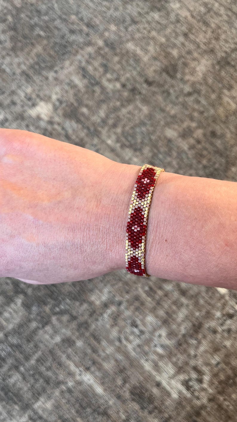 May include: A beaded bracelet with a repeating pattern of red and cream-colored beads. The bracelet is worn on a wrist and has a gold-colored band. The beads are small and tightly woven together.