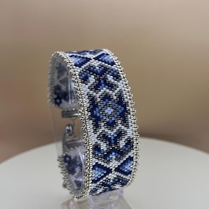 Op de afbeelding: Een kralenarmband met een geometrisch patroon in blauw- en wittinten. De armband wordt gepresenteerd op een transparante standaard, waarbij het gedetailleerde kralenwerk en de zilverkleurige randen worden benadrukt. De sluiting is zichtbaar en het ontwerp is symmetrisch.
