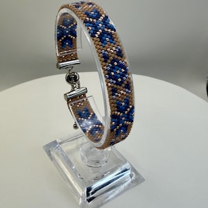 May include: A beaded bracelet with a geometric pattern in shades of blue and tan. The bracelet is displayed on a clear acrylic stand, showcasing its intricate design and silver-toned clasp.