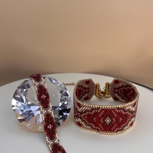 May include: Two beaded bracelets. One is a wide cuff, the other is a narrower band. Both feature a red and white geometric pattern with gold-colored clasps. The bracelets are displayed on a white surface.
