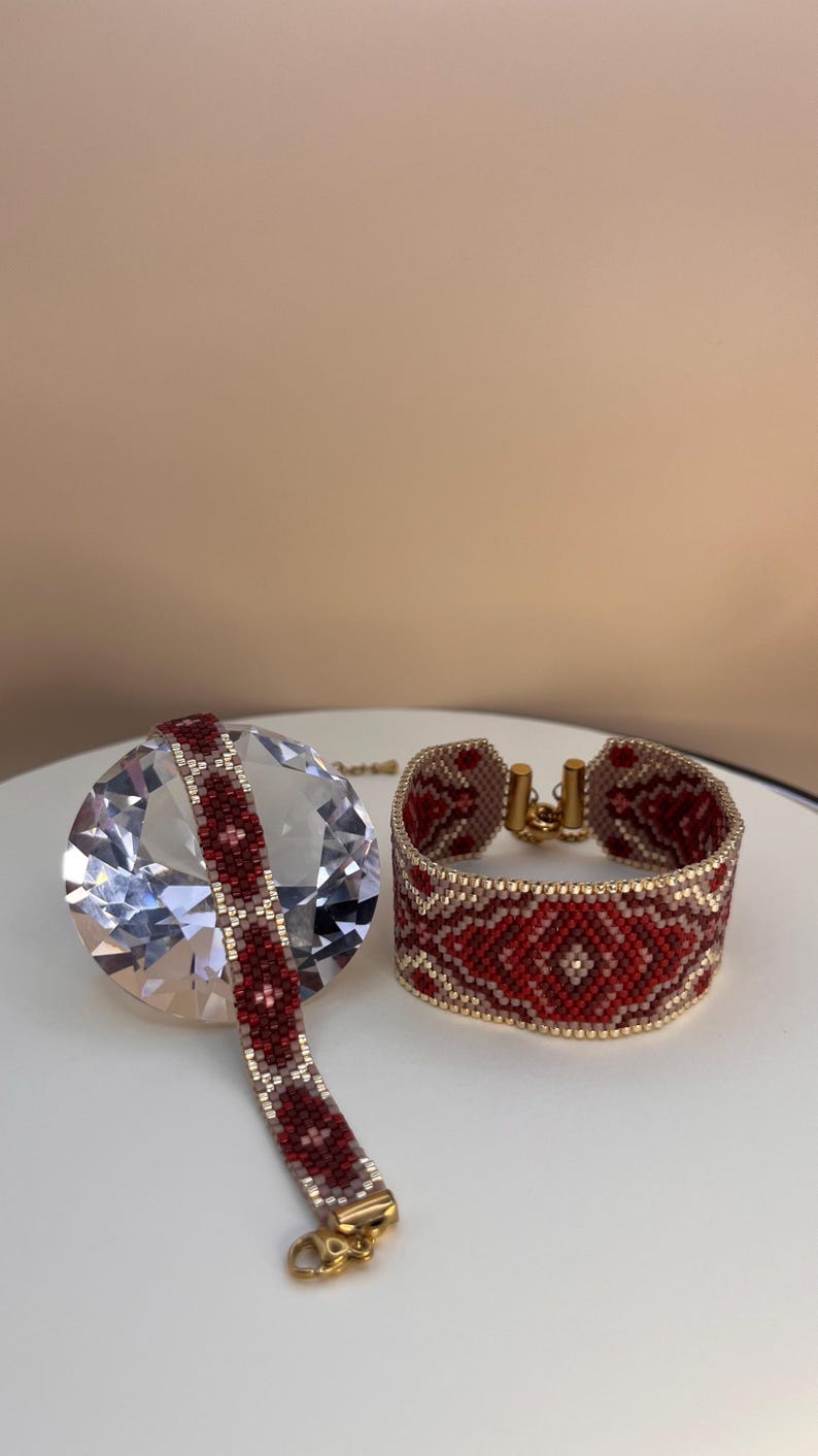 May include: Two beaded bracelets. One is a wide cuff, the other is a narrower band. Both feature a red and white geometric pattern with gold-colored clasps. The bracelets are displayed on a white surface.