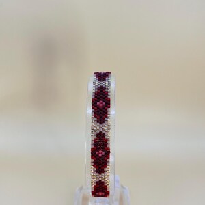 May include: A beaded bracelet with a geometric pattern in shades of red, burgundy, and silver. The bracelet is displayed on a clear stand, showcasing its intricate design. The beads are small and tightly woven, creating a textured surface.