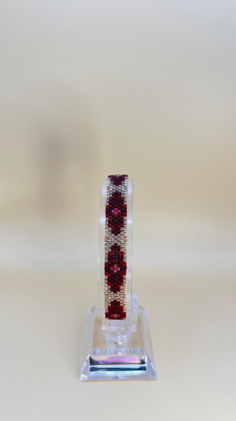 May include: A beaded bracelet with a geometric pattern in shades of red, burgundy, and silver. The bracelet is displayed on a clear stand, showcasing its intricate design. The beads are small and tightly woven, creating a textured surface.