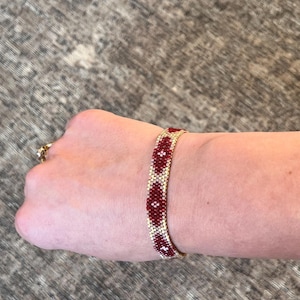 May include: A beaded bracelet with a gold-colored base and a pattern of red and white floral designs. The bracelet is shown on a wrist, showcasing the intricate beadwork and color combination. The bracelet is approximately 0.75 inches wide.