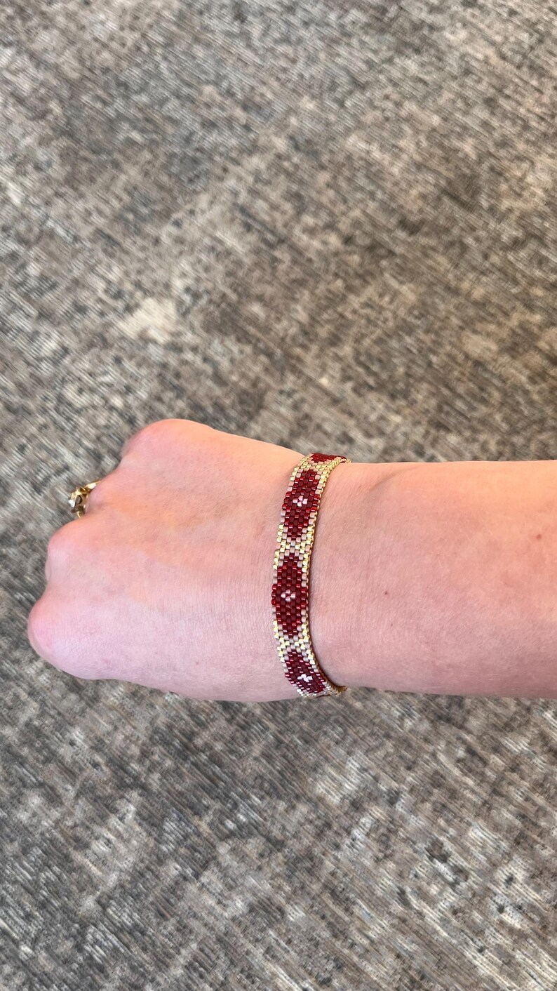 May include: A beaded bracelet with a gold-colored base and a pattern of red and white floral designs. The bracelet is shown on a wrist, showcasing the intricate beadwork and color combination. The bracelet is approximately 0.75 inches wide.